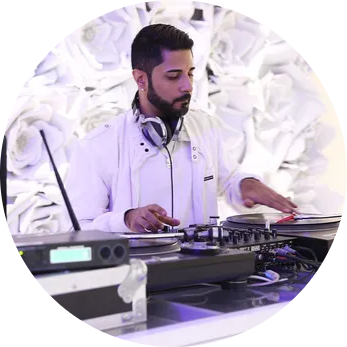 Mixmaster Jash in a white outfit mixes music at a turntable in a stylish setting.