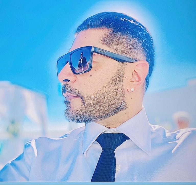 Man with a well-groomed beard, sunglasses, and a formal shirt, set against a bright background.